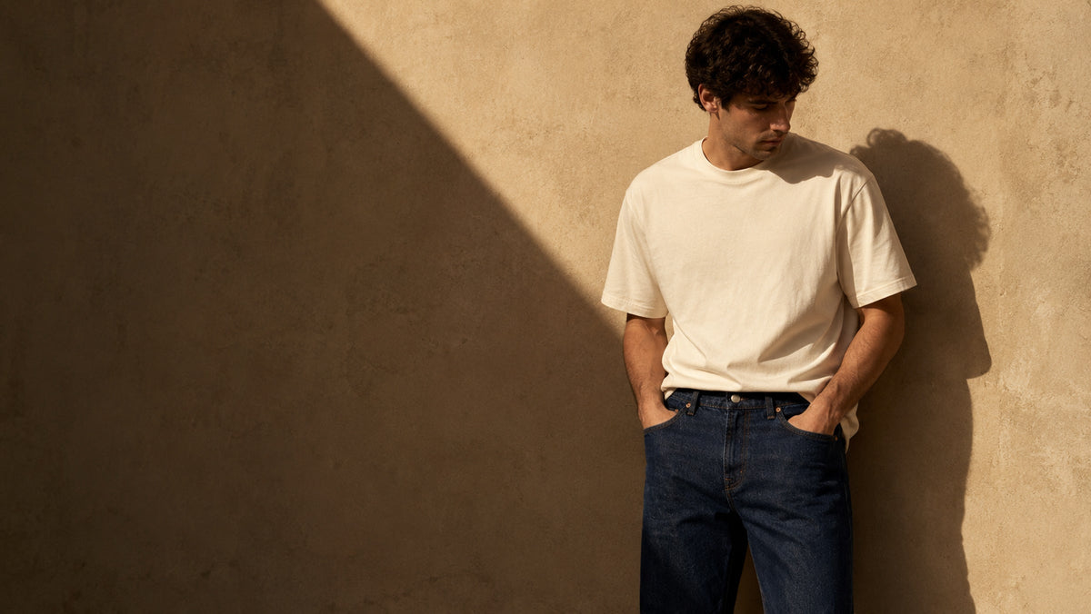 A man leaning against a sunlit sand-coloured wall in an ecru cotton t-shirt and indigo straight leg jeans